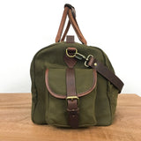 A White Cap Waxed Canvas Duffle from Rogue Industries in green, featuring brown leather accents, a front pocket with a buckle closure, and a full-grain leather shoulder strap, sits on a wooden surface against a plain white background. The water-resistant waxed canvas ensures durability and style.