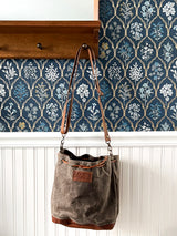 A Rogue Industries Kennebunkport Bucket Bag with an adjustable shoulder strap is hanging on a wall next to a blue wallpaper.