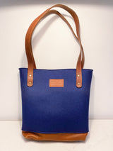 The Sheepscot Felt Wool Tote Bag by Rogue Industries showcases an eye-catching blue design complemented by brown leather straps and a durable leather bottom. Its felted wool construction brings an added charm, highlighted by the distinctive "Rogue Industries" label.