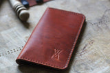 A WM Leatherwork Collection - Premium Leather Field Notes Wallet from Rogue Industries, crafted from brown leather with red stitching and an embossed 'W' logo, lies on a wooden surface next to a blurred object.