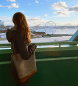 Standing on a balcony and overlooking a waterfront with a Ferris wheel and cranes under a partly cloudy sky, a person with long hair carries the Rogue Industries Sheepscot Felt Wool Tote Bag, known for its lightweight felted wool and leather trim.