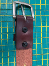 Close-up of a handcrafted Baxter Leather Belt - 1.5" Wide by Rogue Industries made from premium cowhide, featuring a silver buckle secured with two black screws on a green and yellow grid-patterned cutting mat. Made in the USA.